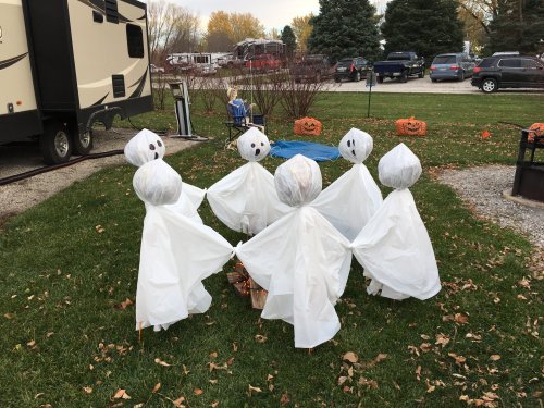 A group of ghost decorations made from white sheets, gathered in a circle on a grassy area, with pumpkins in the background.