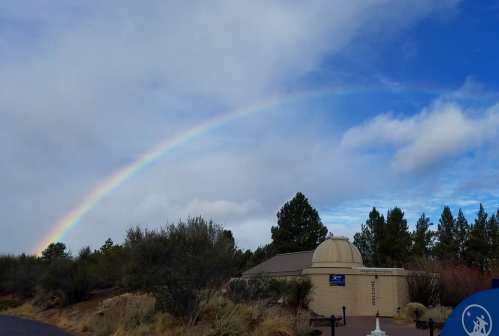 A rainbow arcs over a small building surrounded by trees and shrubs under a partly cloudy sky.