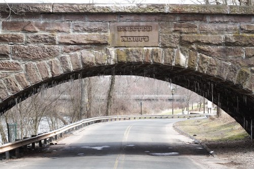 Stone bridge arch with "Erected A.D. 1867" inscription, overlooking a road and trees in a winter landscape.