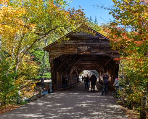 A wooden covered bridge surrounded by vibrant autumn foliage and people walking along the path.