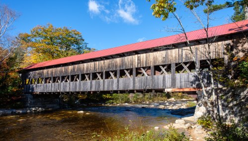 A wooden covered bridge with a red roof spans a river, surrounded by autumn foliage and a clear blue sky.