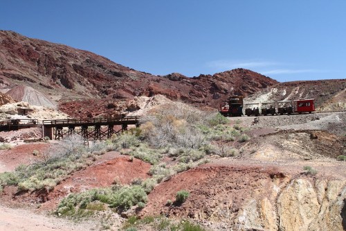 A scenic view of colorful rocky hills with a train on a bridge in a desert landscape under a clear blue sky.