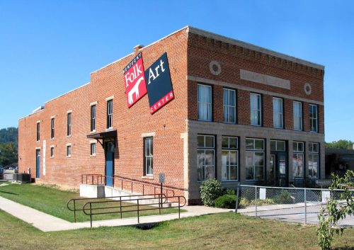 Exterior view of the Folk Art Center, a brick building with large windows and a prominent sign, surrounded by grass.