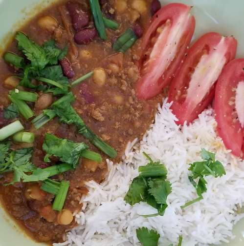 A bowl of chili with beans, topped with fresh herbs, served alongside rice and sliced tomatoes.
