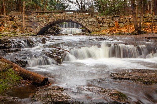 A serene landscape featuring a stone bridge over a flowing waterfall, surrounded by trees and rocky terrain.