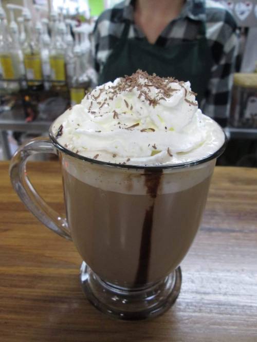 A glass of hot chocolate topped with whipped cream and chocolate shavings, set on a wooden counter.