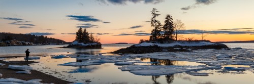 A serene sunset over a frozen lake, with ice chunks and a person standing on the shore near small islands.
