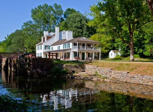 Historic white house with green shutters by a calm river, surrounded by trees and a stone wall. Clear blue sky above.