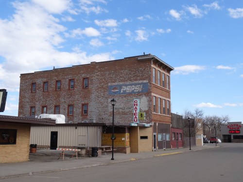 A weathered brick building with a faded Pepsi sign, located on a quiet street under a blue sky with scattered clouds.