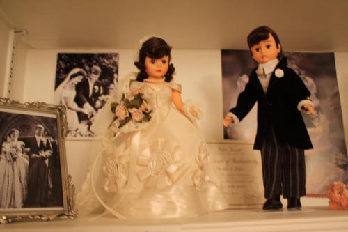 Two wedding dolls on a shelf: a girl in a white dress and a boy in a tuxedo, with wedding photos in the background.
