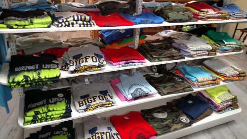 A display of colorful t-shirts stacked on shelves, featuring various designs and slogans related to Bigfoot.