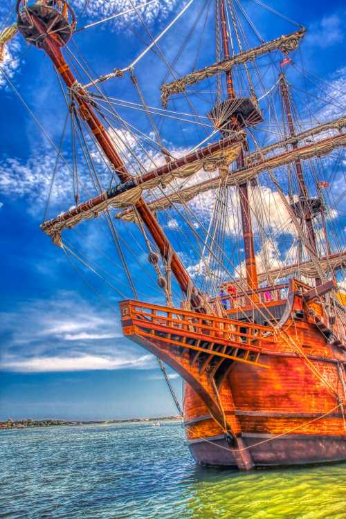 A majestic wooden ship with tall masts and sails, anchored in calm waters under a bright blue sky.