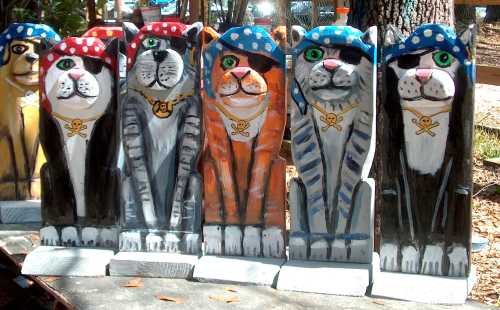 Colorful wooden pirate cat sculptures, each wearing a bandana and eye patch, lined up in a row.