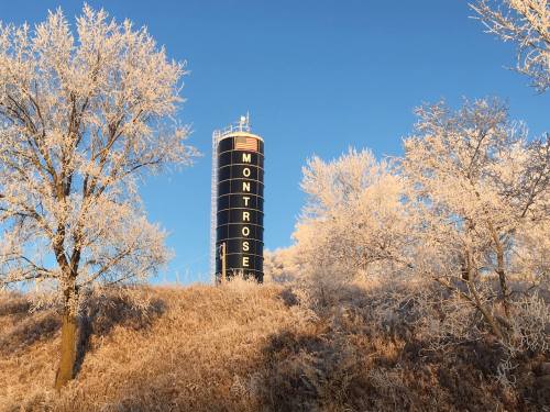 A black water tower labeled "Montrose" stands amidst frosty trees and grass under a clear blue sky.