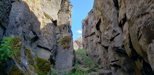 A narrow rocky canyon with tall stone walls and green grass, under a clear blue sky.
