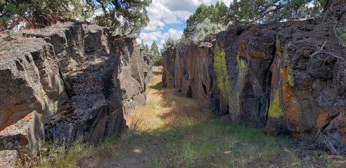 A narrow pathway between tall, rugged rock formations, surrounded by grass and trees under a partly cloudy sky.