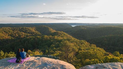 A couple sits on a rock overlooking a lush green valley at sunset, with a serene lake in the distance.