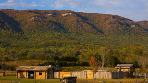 A scenic view of rustic log cabins in a field, with colorful autumn mountains in the background under a blue sky.