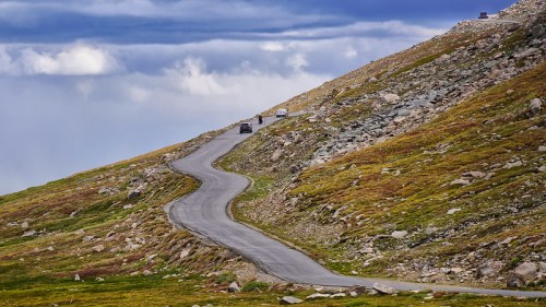 Winding road through rocky terrain and grassy hills under a cloudy sky. Vehicles are seen traveling along the road.