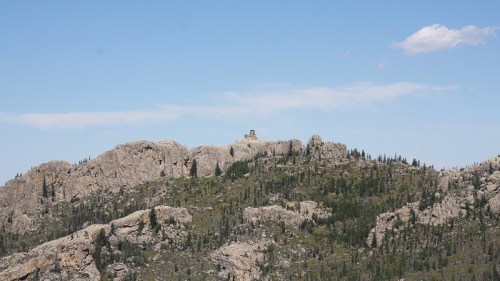 A distant view of a rocky mountain landscape with a small structure atop a peak under a clear blue sky.