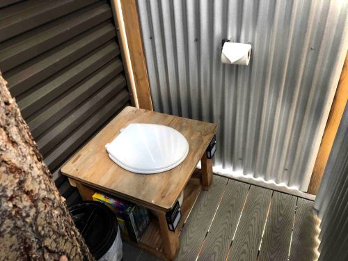 A rustic outdoor toilet setup with a wooden seat, toilet paper, and a tree nearby, enclosed by corrugated metal walls.