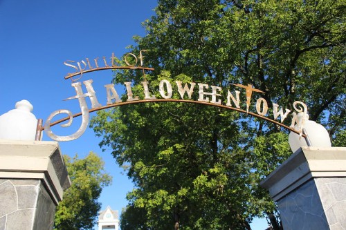 Sign reading "Spirit of Halloweentown" arching over a pathway, surrounded by trees and a clear blue sky.