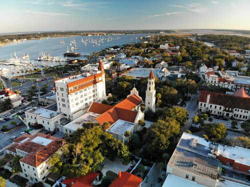 Aerial view of a coastal town with marinas, historic buildings, and lush greenery under a clear sky.