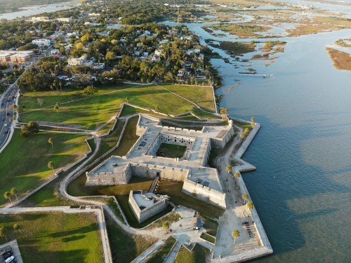 Aerial view of a historic fort surrounded by water and greenery, with a nearby town in the background.