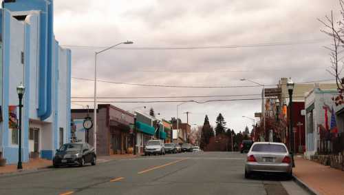 A quiet street scene featuring storefronts, parked cars, and overcast skies in a small town.