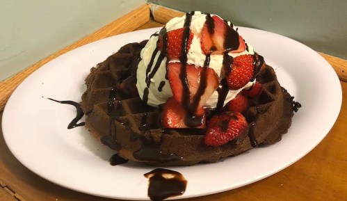 A chocolate waffle topped with whipped cream, fresh strawberries, and drizzled with chocolate sauce on a white plate.
