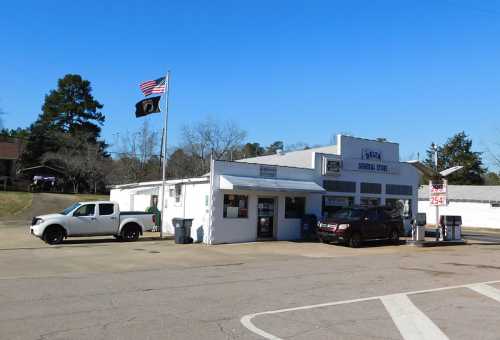 A small general store with a flag, parked vehicles, and trees in the background under a clear blue sky.