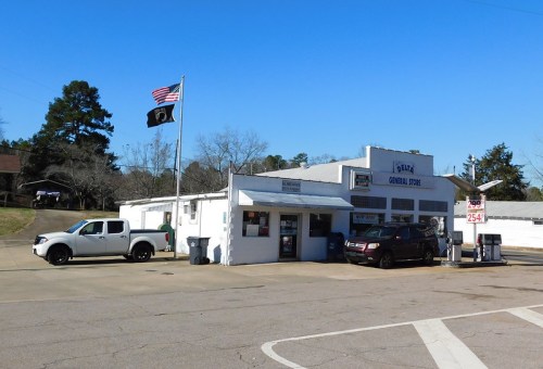 A small general store with a flag, parked vehicles, and trees in the background under a clear blue sky.