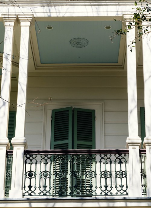A close-up of a balcony with green shutters and an ornate iron railing, framed by white columns and a blue ceiling.