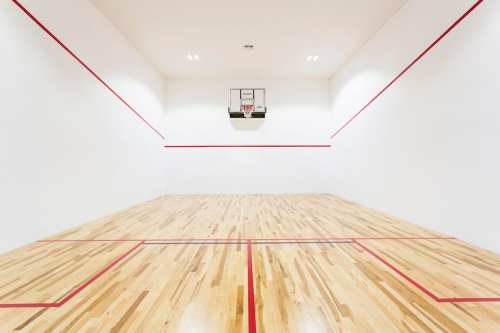A spacious indoor squash court with wooden flooring and red lines, featuring a wall-mounted hoop at one end.