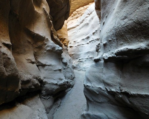 Narrow canyon with smooth, layered rock walls and a sandy floor, illuminated by light from above.