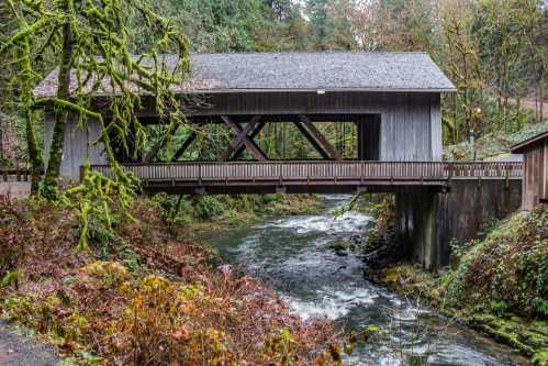 A wooden covered bridge spans a flowing river, surrounded by lush greenery and autumn foliage.