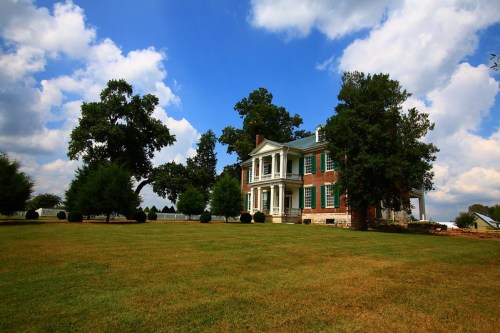 A historic two-story brick house with green shutters, surrounded by trees and a grassy lawn under a blue sky with clouds.