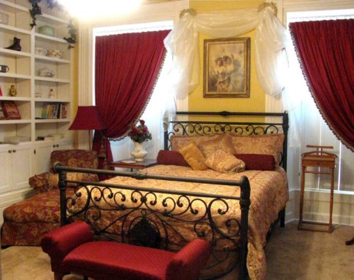 Cozy bedroom featuring a wrought iron bed, red curtains, decorative pillows, and a bookshelf in a warm, inviting atmosphere.