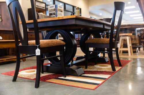 A wooden dining table with a unique base, surrounded by chairs on a patterned rug in a furniture store.