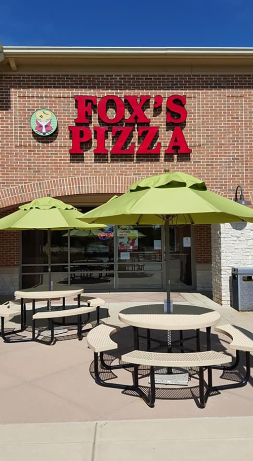 Exterior of Fox's Pizza with green umbrellas over picnic tables in front of a brick building.