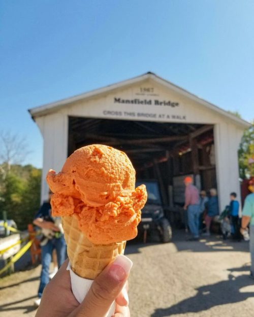 A hand holds a cone with orange ice cream in front of a covered bridge and people enjoying a sunny day.