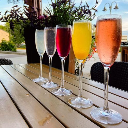 A row of six colorful cocktails in tall glasses on a wooden table, with flowers and outdoor seating in the background.