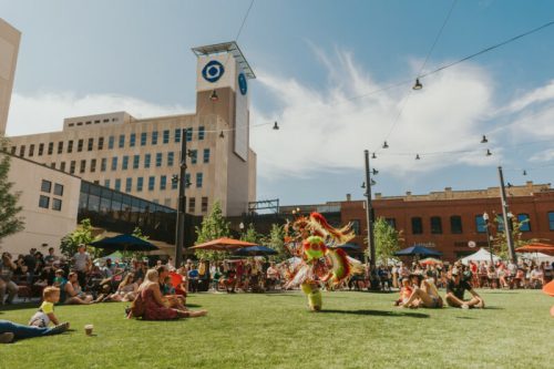 A vibrant outdoor festival scene with a performer in a colorful costume dancing on grass, surrounded by an audience.