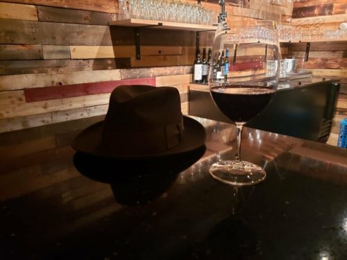 A dark fedora hat rests on a bar counter next to a glass of red wine, with a rustic wooden backdrop.