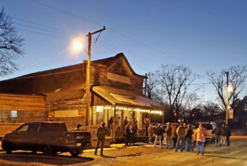 A crowd gathers outside a rustic wooden building at dusk, illuminated by string lights and street lamps.