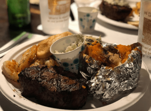 A plate of grilled steak, shrimp, baked potato, and a small cup of coleslaw, served with drinks in the background.