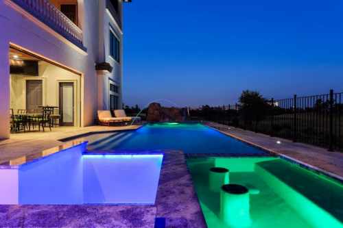 A modern backyard at dusk featuring a pool with colorful lights, lounge chairs, and a patio area.