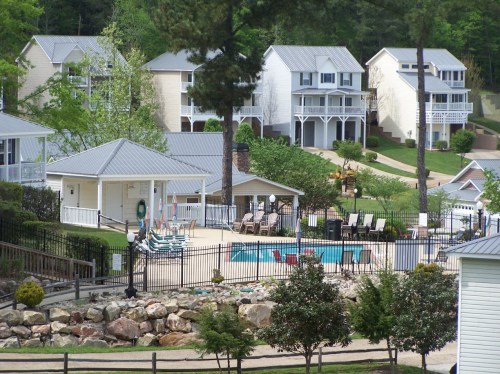 A scenic view of a residential area with a pool, lounge chairs, and nearby houses surrounded by trees and landscaping.