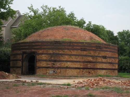 A round, brick structure with a domed roof, partially covered in grass, surrounded by trees and construction debris.
