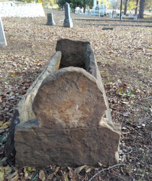 An old, weathered stone grave marker in a cemetery, surrounded by fallen leaves and other gravestones.